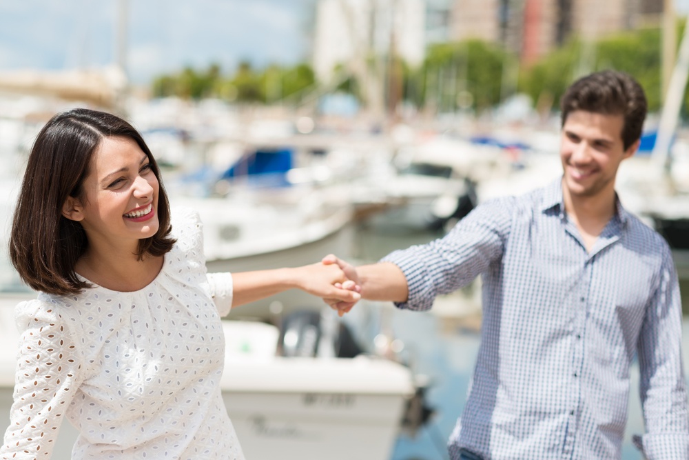 Attractive couple exploring on their summer vacation laughing as they hold hands while sightseeing a small boat harbour on the coast